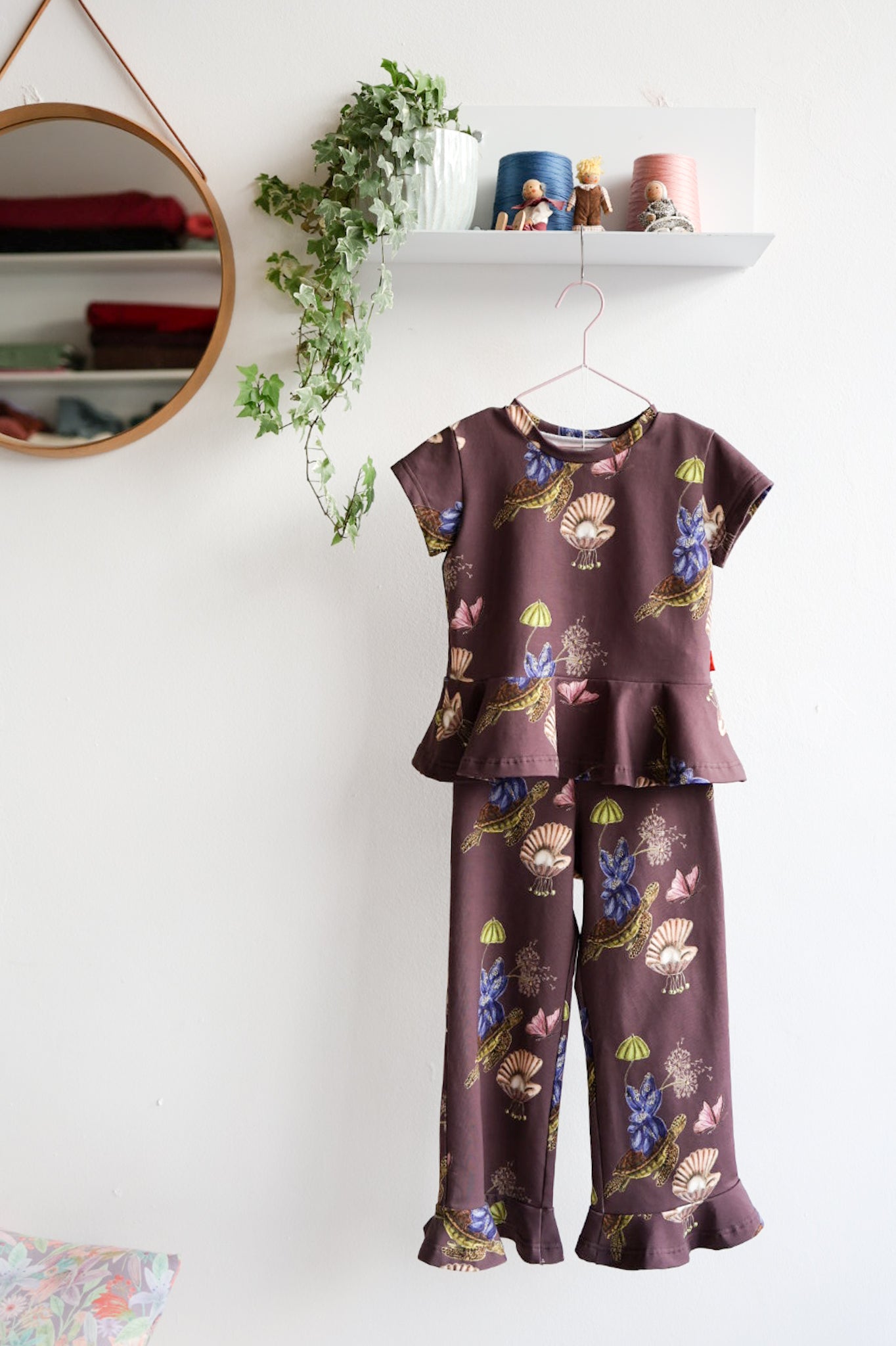 Kids nature-themed print top and pants hanging on a white wall with decorative items.