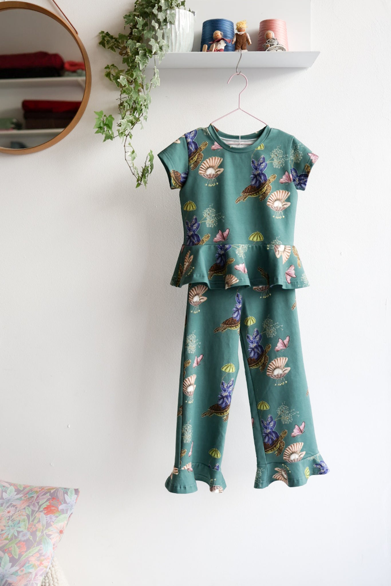 Kids nature-themed print top and pants hanging on a white wall with decorative items.