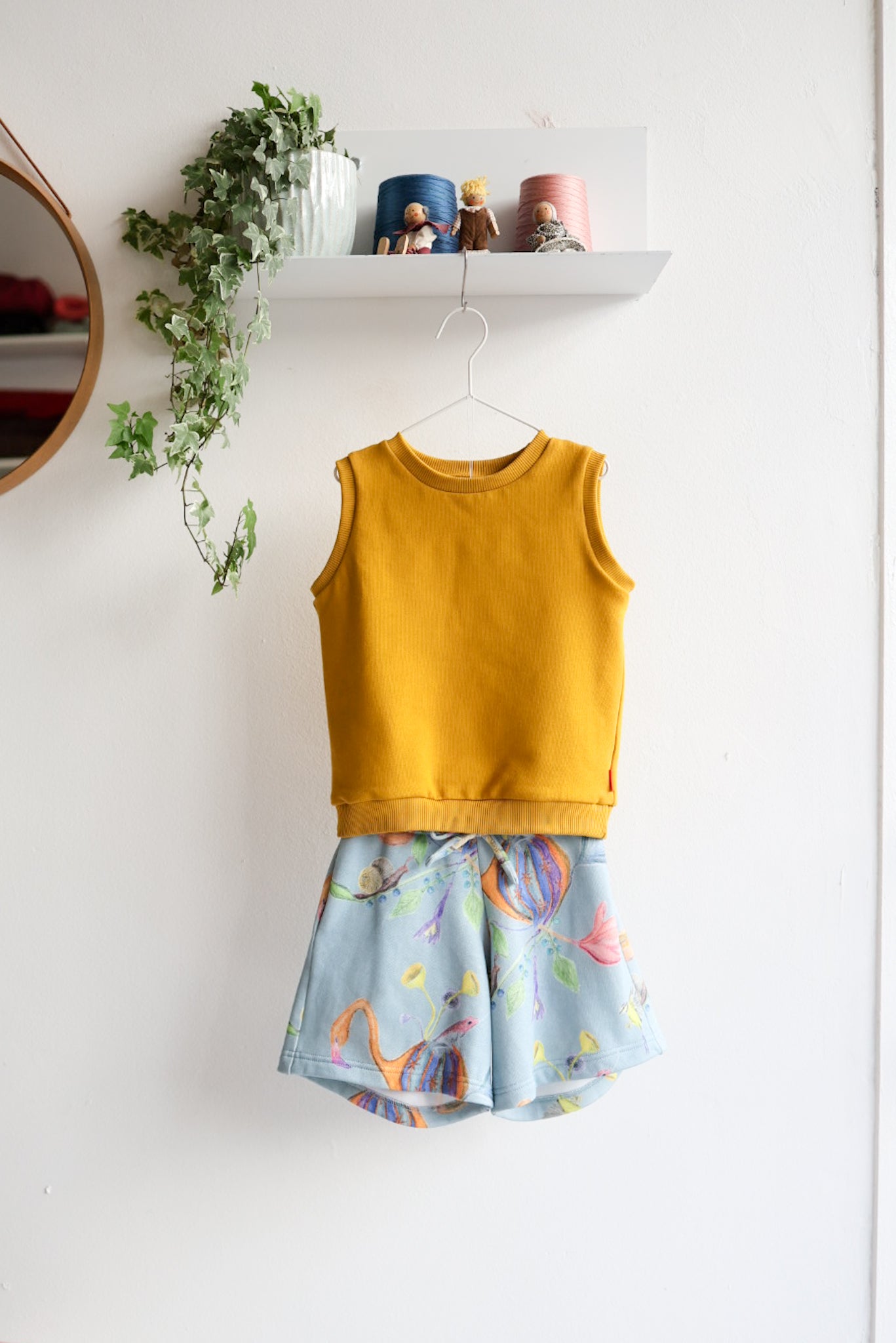 Mustard yellow kids sleeveless top and colorful shorts on a white wall with decorative elements.