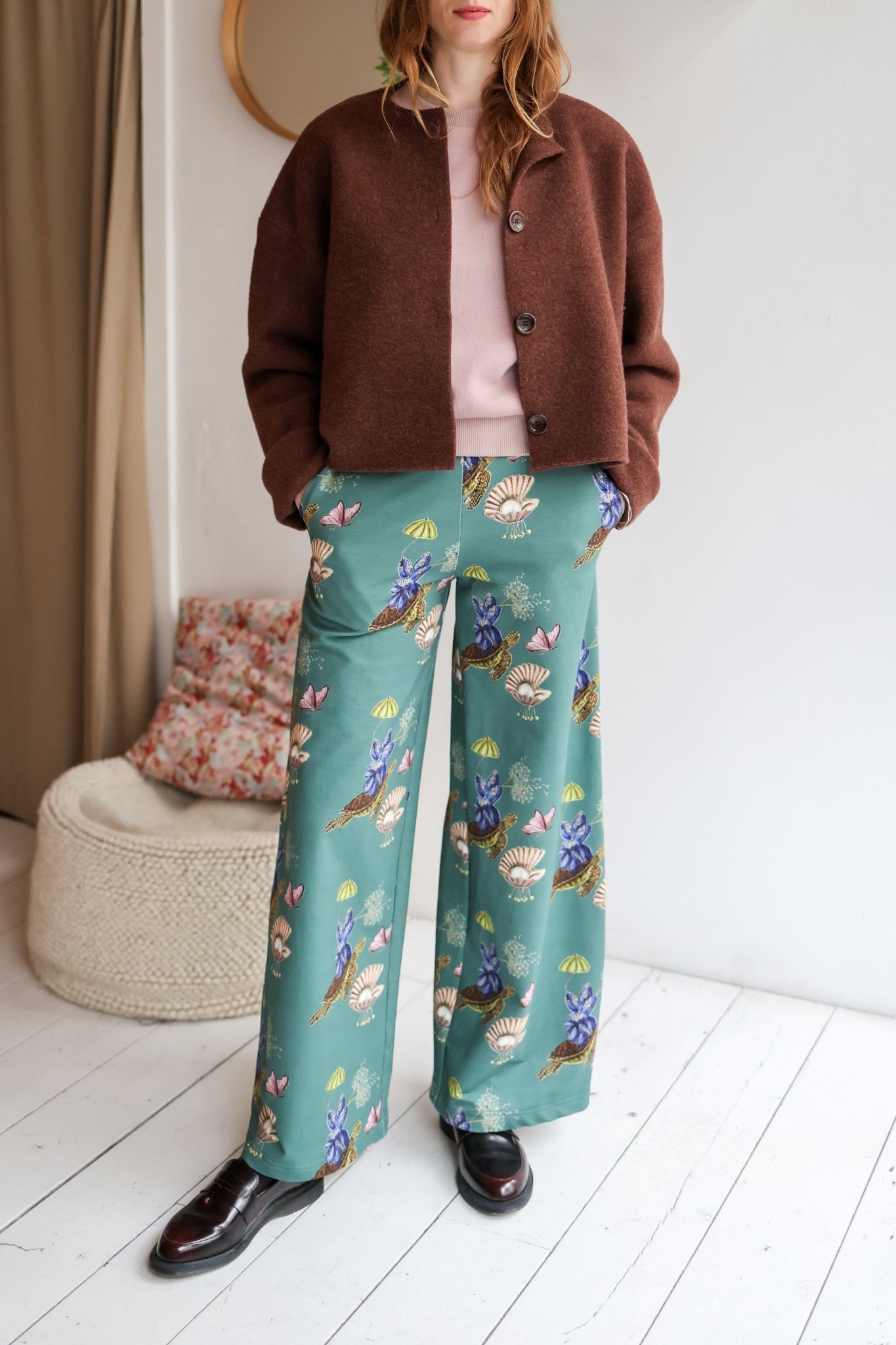 Woman wearing a pink sweater and teal floral pants in a room with a white wall and wooden floor.
