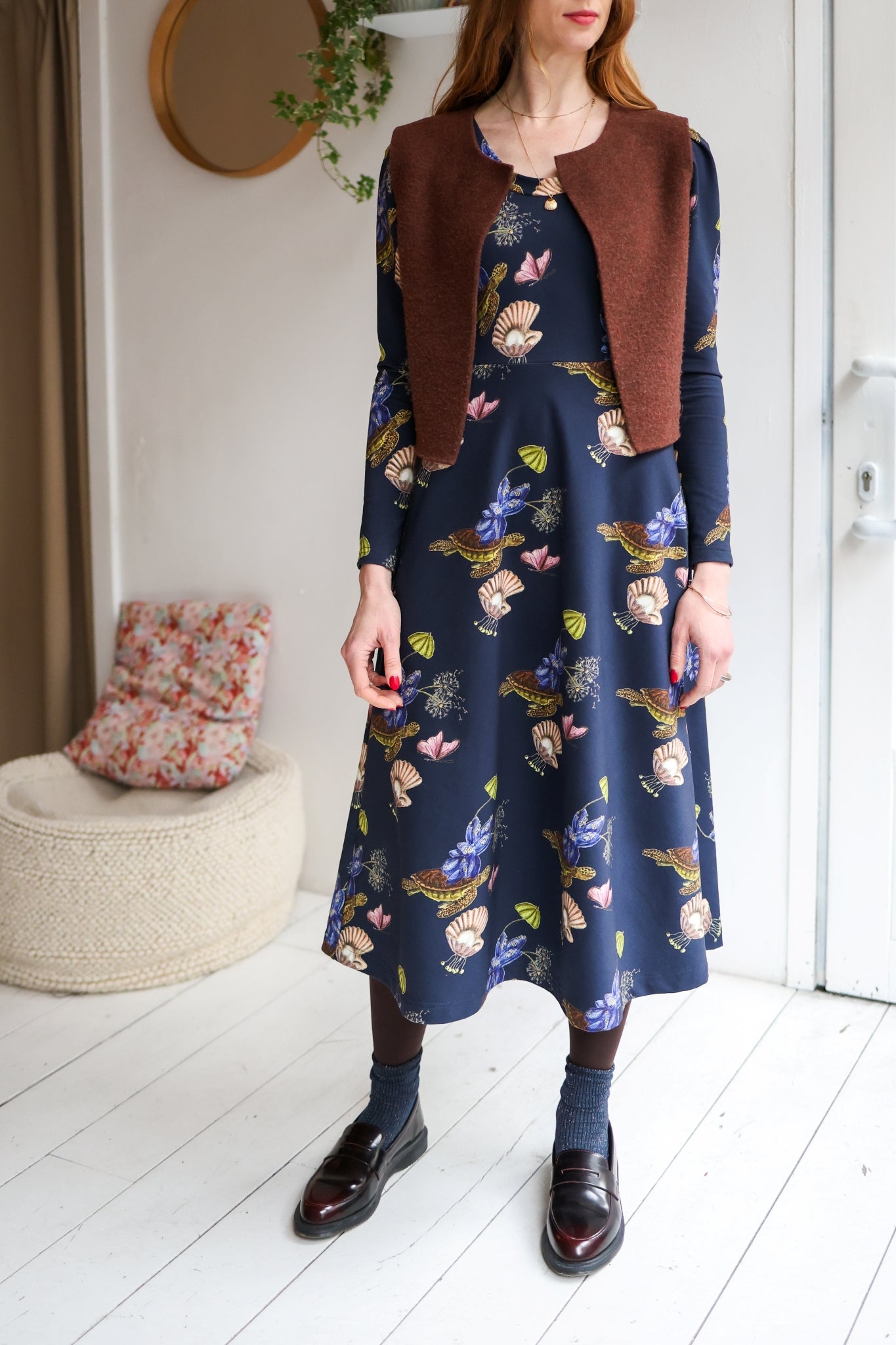 Woman wearing a brown vest over a patterned dress in a room with a white wall.