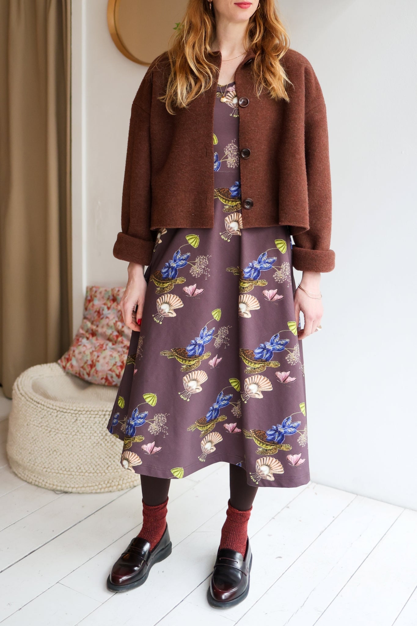 Woman wearing a brown jacket and a patterned dress with long sleeves with dark shoes indoors.