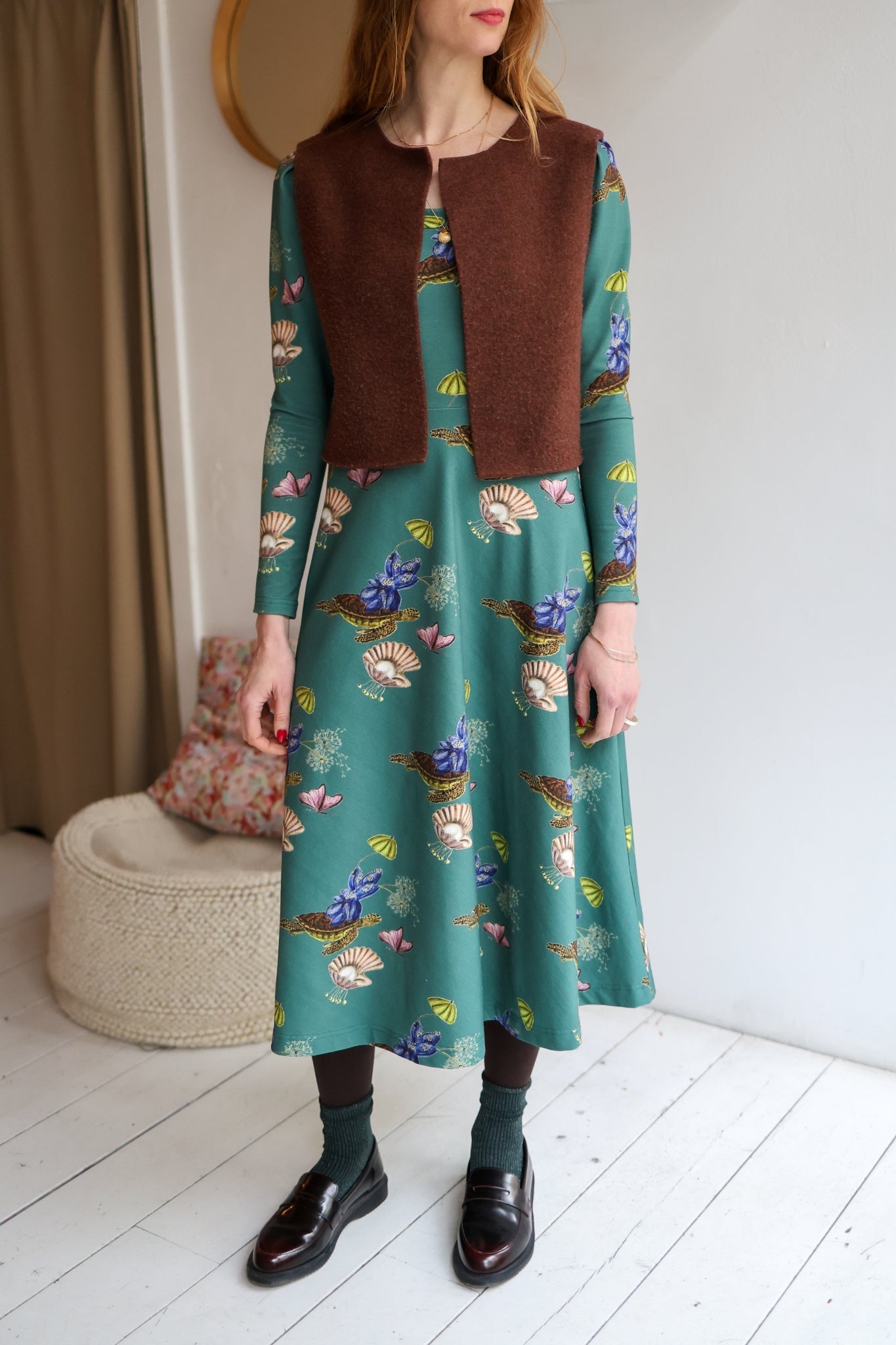 Woman wearing a green patterned dress with a brown vest indoors.