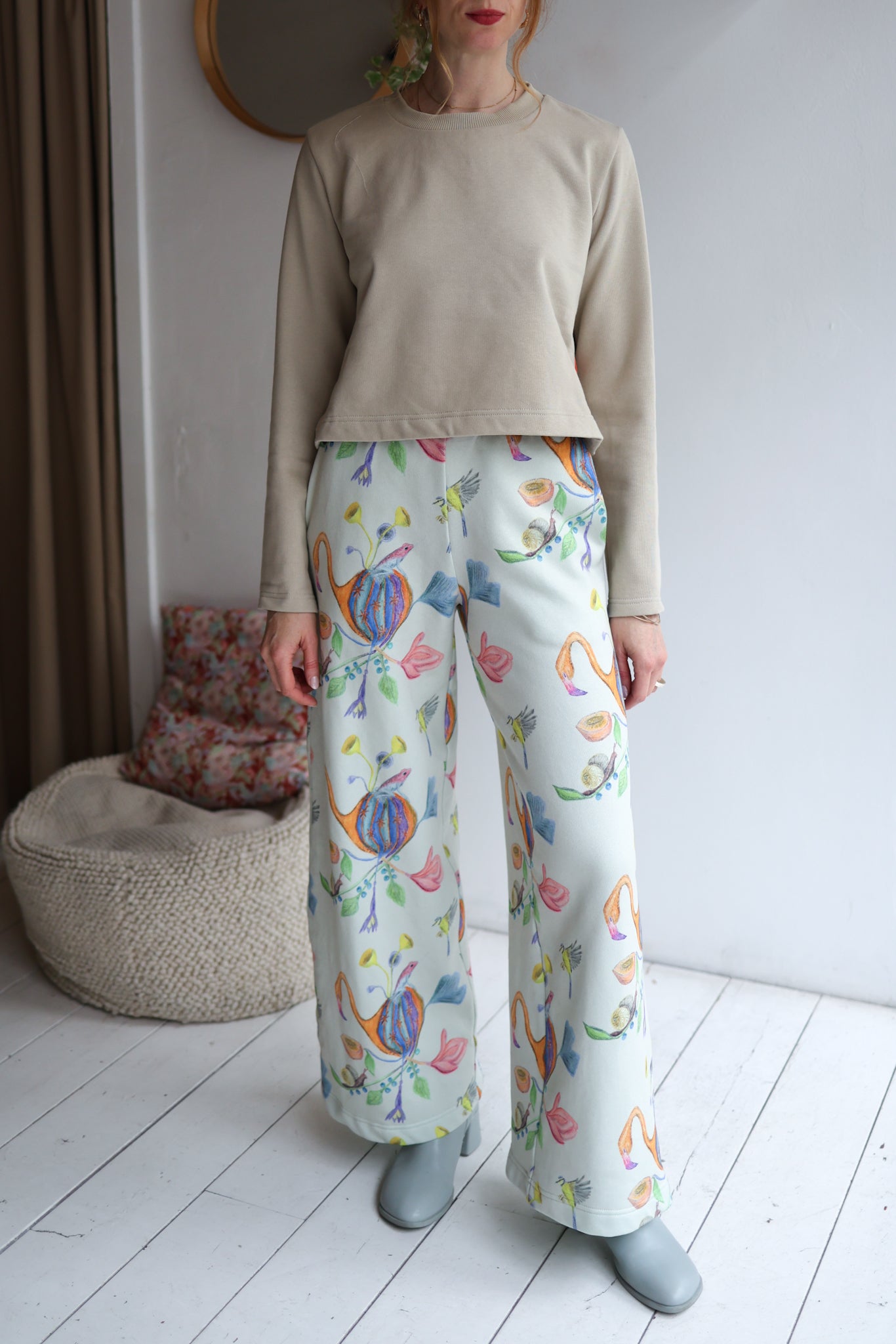 Person wearing a beige sweater and colorful patterned pants standing in a room with a white wall and wooden floor.