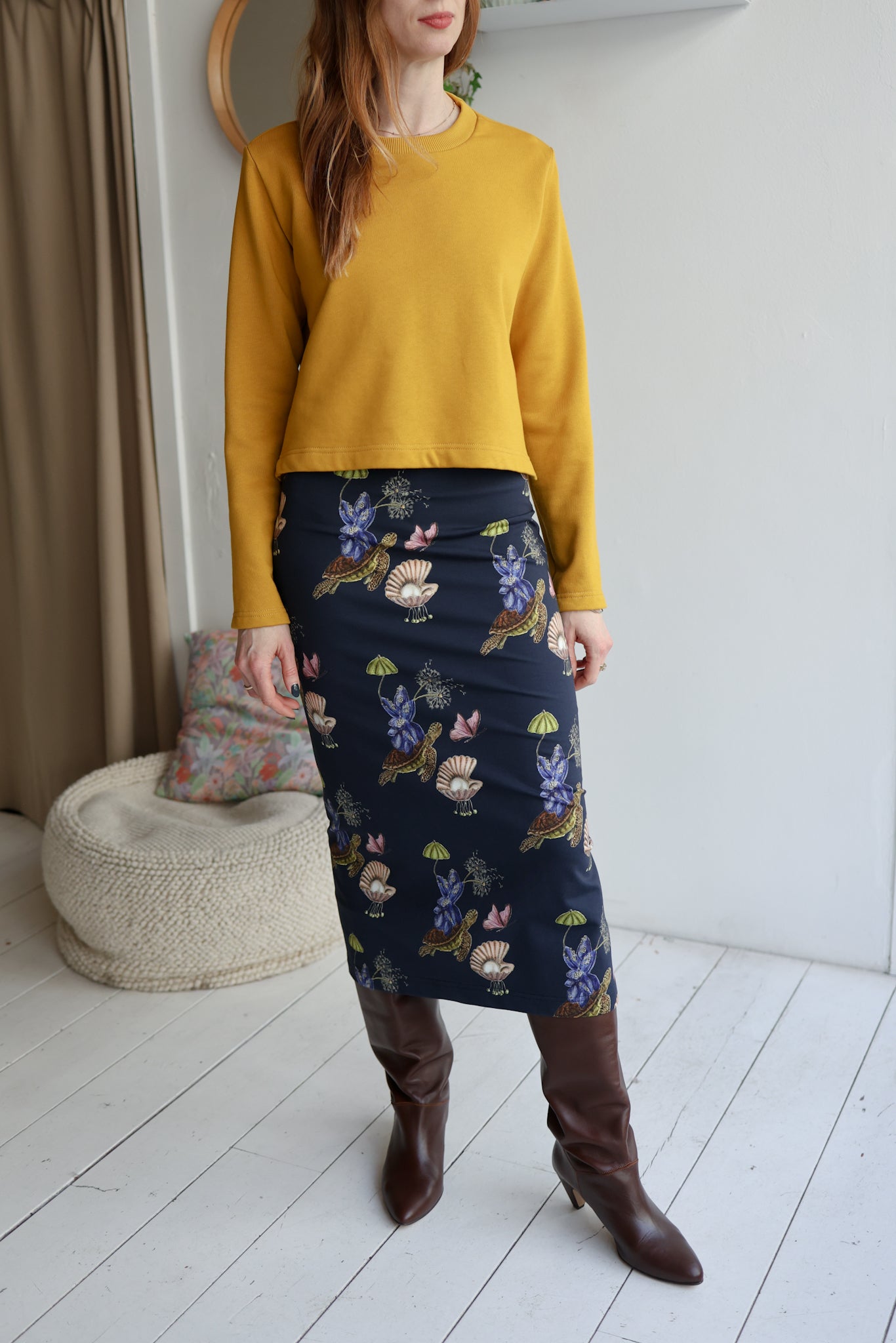 Woman wearing a yellow sweatshirt and a navy patterned skirt in a room with white walls and a wooden floor.